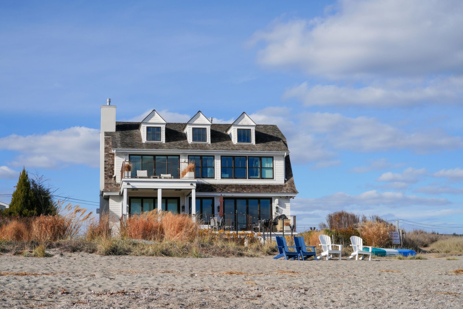Cozy beachfront house with patio chairs surrounded by sand and grasses under blue sky.